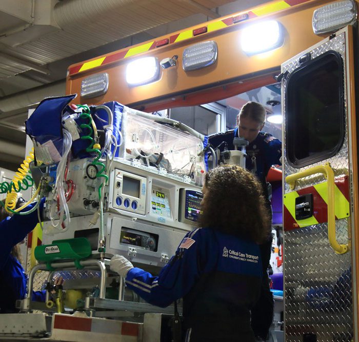 A medical team is loading an advanced patient transport unit into an ambulance. The unit is equipped with various medical devices, including monitors and an incubator. Two personnel, one adjusting equipment inside the ambulance and the other preparing the transport unit, are wearing matching blue uniforms. The ambulance is brightly lit with emergency lights, indicating an urgent scenario.