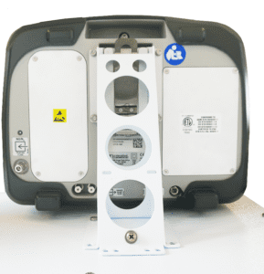 The image shows the back of a device with a mounting bracket. The device has a gray casing with a large circular opening in the center surrounded by three smaller circular cutouts. There are two white panels on the sides, one featuring a caution label and the other various informational labels. Near the top, there is a blue symbol indicating accessibility. The overall design appears modern and technical, suggesting it may be an electronic or monitoring device.