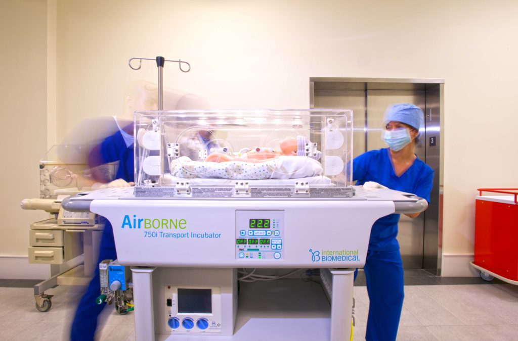 A medical setting featuring a transport incubator labeled "AirBORNE 750i" on a wheeled cart. Inside the incubator, a newborn baby is visible, wrapped in a patterned blanket. Two healthcare professionals in blue scrubs and face masks are depicted; one is pushing the incubator while the other is attending to the baby. The background shows medical equipment and an elevator. The atmosphere is clinical and focused on neonatal care.