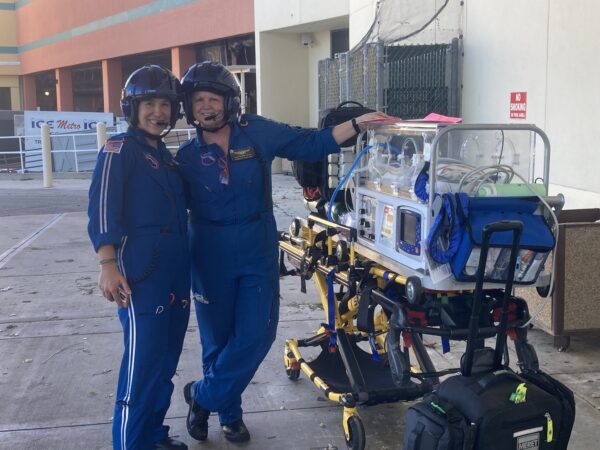 Two women in blue flight suits and helmets smile for the camera. They stand next to a medical transport stretcher with a clear enclosure on it, indicating readiness for patient transport. The background features a light-colored wall and some outdoor seating, suggesting a public space. Both women appear confident and friendly.