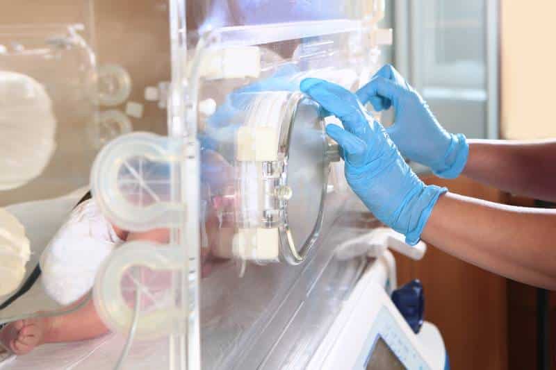 Gloved hands of a doctor working in the NICU closing the door on an incubator with an infant inside.