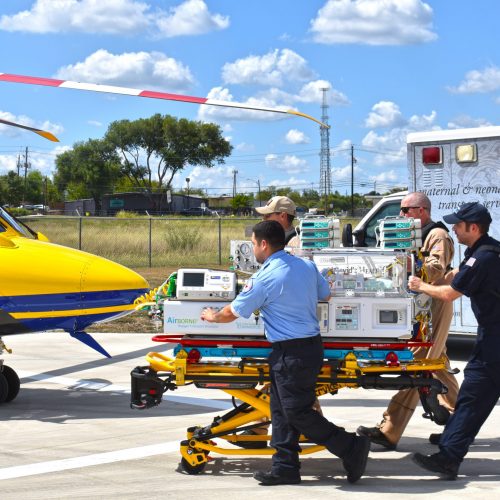 A bright yellow and blue helicopter is parked on a helipad under a partly cloudy sky. In the foreground, a team of four emergency responders is moving a stretcher with medical equipment on it. One responder in a blue uniform is pushing the stretcher, while another, wearing a tan uniform, is walking beside him. A third responder, dressed in a dark uniform with a backpack, is seen further back, and a fourth responder in a tan uniform is assisting with the equipment. In the background, a medical ambulance is parked with its side displaying images related to maternal and neonatal transport services, alongside a green landscape and a fence.