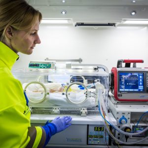 A female medical professional in a bright yellow jacket and blue gloves is attending to a newborn in an incubator. The incubator is equipped with monitoring and medical equipment. To the right, a medical device displays vital signs and other data. The scene is well-lit, emphasizing the attentive care being provided to the infant.